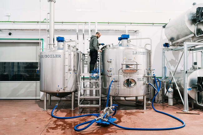 Seitenansicht eines männlichen Ingenieurs bei der Untersuchung eines Edelstahltanks mit Schläuchen in einer Brauerei mit feuchtem Boden — Stockfoto