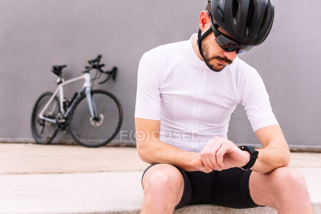 Male biker in protective helmet and sunglasses watching heart rate on wearable bracelet while sitting against bicycle on three color background - foto de stock