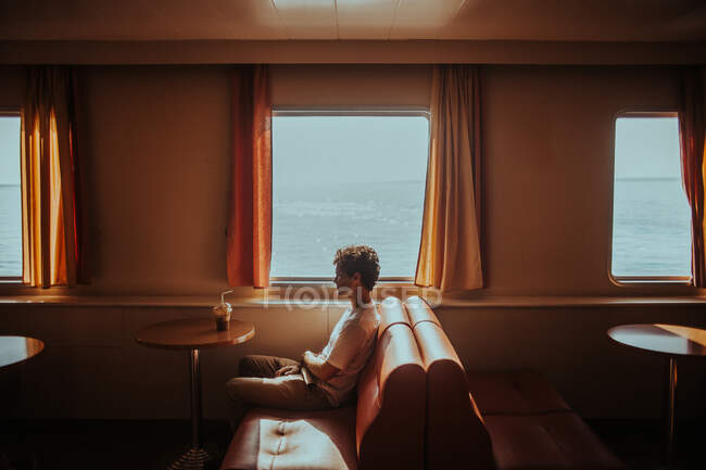 Vue latérale réfléchi jeune homme portant des vêtements d'été décontractés assis sur un canapé doux tout en voyageant en ferry sur la mer ondulante en Grèce lumineux — Photo de stock