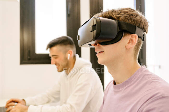 Seitenansicht positiver junger männlicher Angestellter in Freizeitkleidung, der Virtual Reality im modernen Headset erlebt, während er mit einem lächelnden ethnischen Kollegen am Laptop arbeitet — Stockfoto