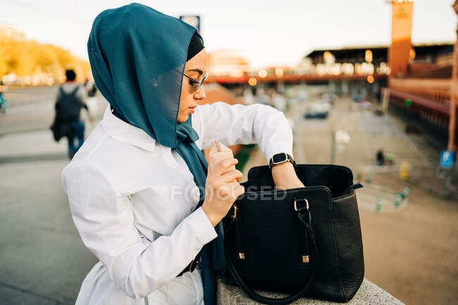 Вид збоку на стильну етнічну жінку в хіджабі і модні сонцезахисні окуляри, що стоять на вулиці і виймають речі з шкіряної сумки — стокове фото