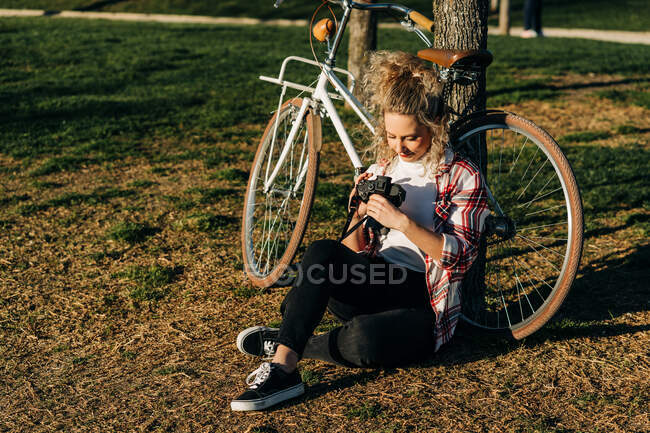 Conteúdo fêmea sentada perto da bicicleta no jardim e olhando através de fotos na câmera fotográfica profissional — Fotografia de Stock