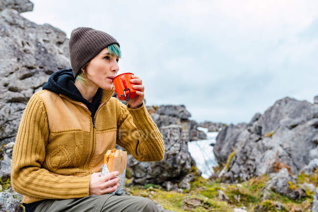 Young female with dyed hair wearing warm clothes and having hot drink with sandwich while sitting on rock in meadow of Peaks of Europe — Stock Photo