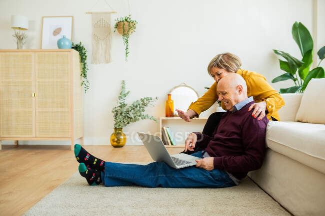 Веселая зрелая пара разговаривает в видеочате на ноутбуке и машет руками в гостиной — стоковое фото