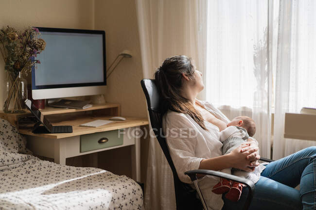 Mamá adulta en ropa casual amamantando a un niño pequeño y encantador mientras está sentada en la habitación de la casa ligera - foto de stock