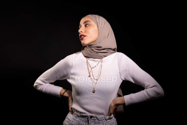 Модиш уверенной мусульманской женщины в хиджабе стоя и глядя в сторону в темную студию — стоковое фото