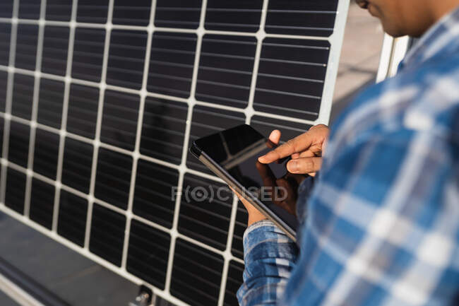 Cropped unrecognizable ethnic male technician in checkered shirt browsing tablet while standing near photovoltaic panel located in modern solar power farm — Stock Photo