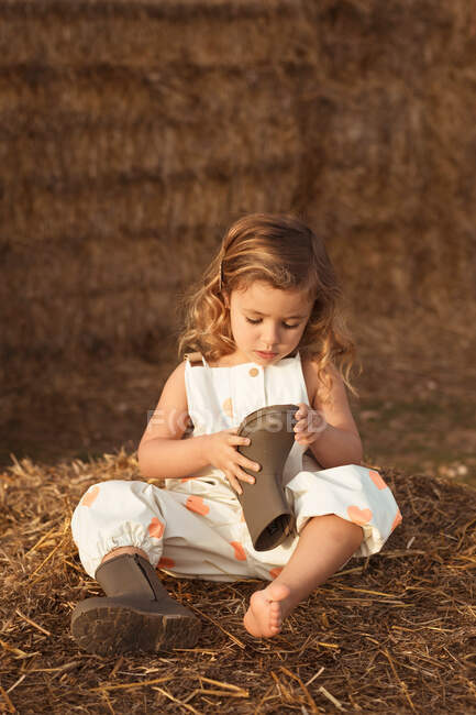 Adorable curious child in overalls sitting on haystack and looking at boots in evening in countryside — Stock Photo