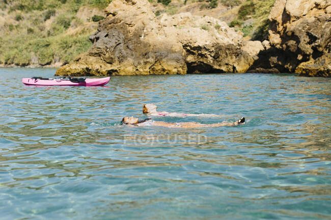Расслабленные подруги, мирно плавающие на теплой лазурной морской воде возле плавающего розового каяка в солнечный день в Малаге Испания — стоковое фото