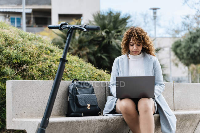 Focused young African American female in blue coat working on netbook while sitting on stone bench near scooter in city park on clear spring day — Fotografia de Stock