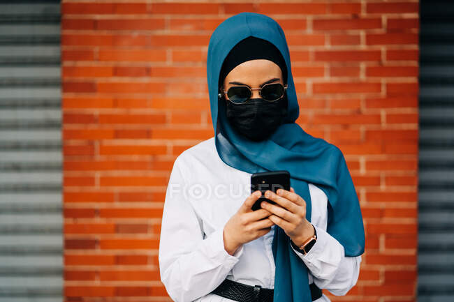 Muslim female wearing protective mask and traditional headscarf standing against wall in city and browsing phone — Photo de stock