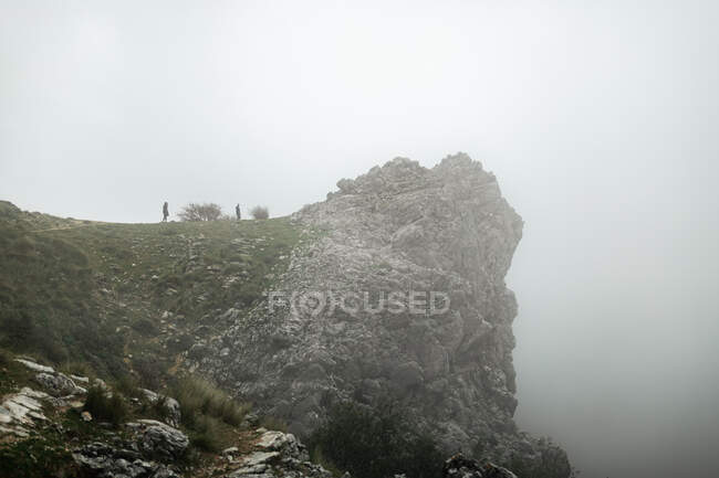 Senderistas distantes parados en la áspera cumbre rocosa de la montaña y admirando los paisajes de las tierras altas bajo el cielo nublado y sombrío - foto de stock