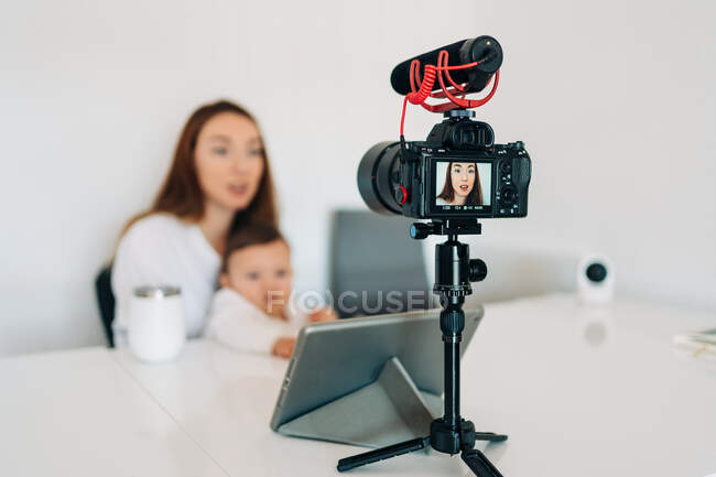 Verschwommene junge Mutter mit süßem Baby auf dem Schoß spricht und nimmt Video für persönlichen Blog auf, während sie zu Hause am Schreibtisch sitzt — Stockfoto