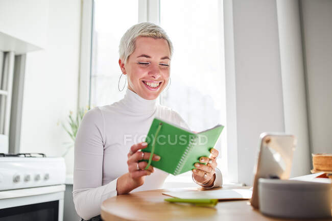 Adult female astrologist reading notepad at desk with smartphone and books at home in sunlight — Stock Photo