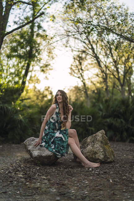 Ganzkörperzufriedene junge Frau trägt trendige Maxi-Sundress, die die Haare sanft berührt und lächelnd wegschaut, während sie auf einem großen Stein im grünen, sonnigen Wald sitzt — Stockfoto