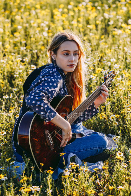 Mujer hipster joven pensativo sentado en un prado en el campo tocando la guitarra durante la luz del sol de verano - foto de stock
