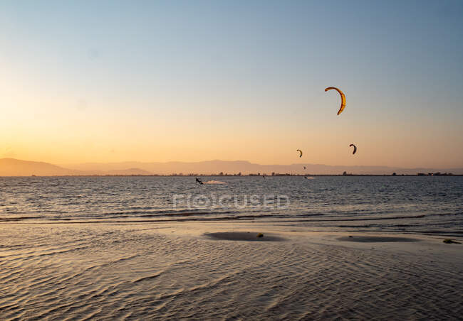 Distant unrecognizable adventurers paragliding over calm rippled sea water against sunset sky in summer evening - foto de stock