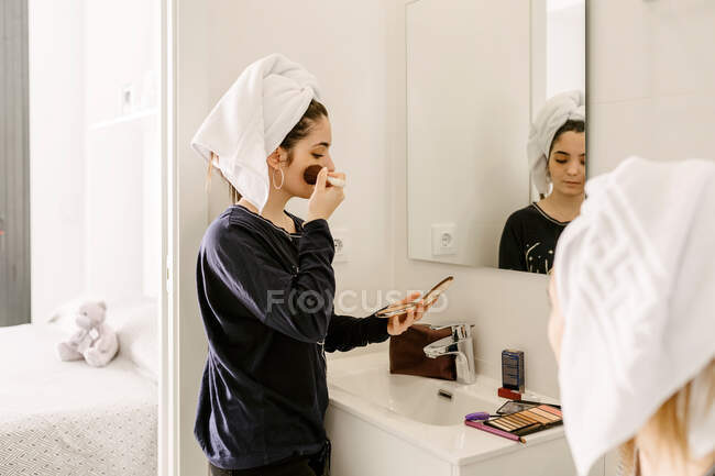 Side view of young Hispanic lady in casual clothes and towel on head applying foundation on face with brush while standing in front of mirror near unrecognizable female friend — Photo de stock