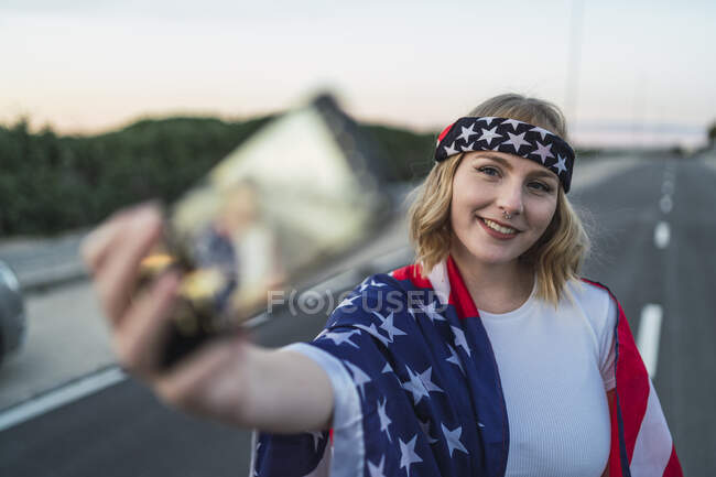 Patriótica mujer americana envuelta en la bandera nacional de EE.UU. tomando selfie en el teléfono móvil mientras está de pie en la carretera - foto de stock