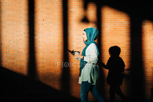 Elegante mujer musulmana en hijab caminando con café para llevar en la estación de tren y navegar por Internet en el teléfono móvil - foto de stock