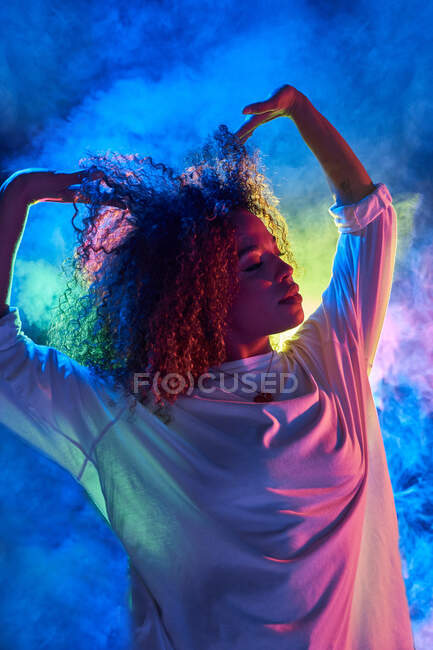 Portrait of attractive young African American female in white shirt touching curly hair while dancing in neon lights in dark studio — Stock Photo
