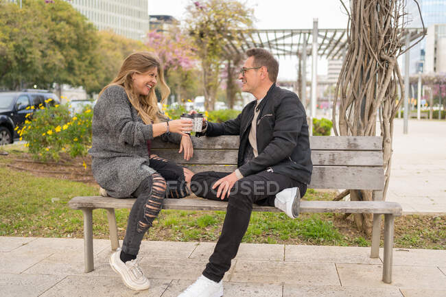 Seitenansicht eines stilvollen Paares mittleren Alters, das auf einer Bank in der Straße sitzt und Kaffee to go trinkt, während man sich gegenseitig ansieht — Stockfoto
