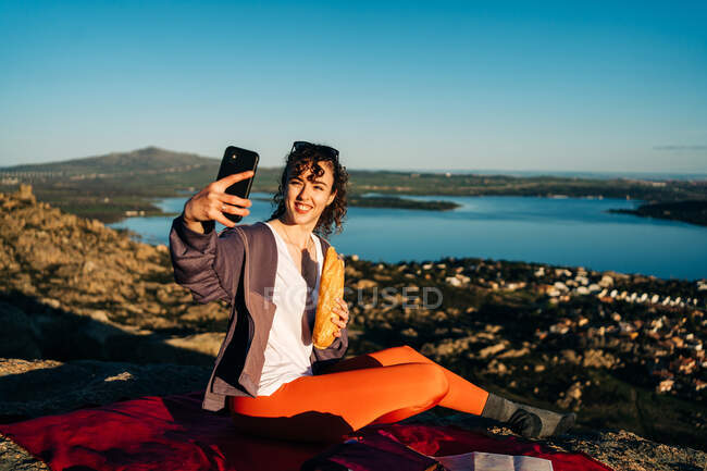 Vista lateral de jovens caminhantes felizes em roupas casuais comendo sanduíche e tomando selfie com telefone celular enquanto sentado em cobertor na colina rochosa sobre o mar contra o céu azul sem nuvens — Fotografia de Stock