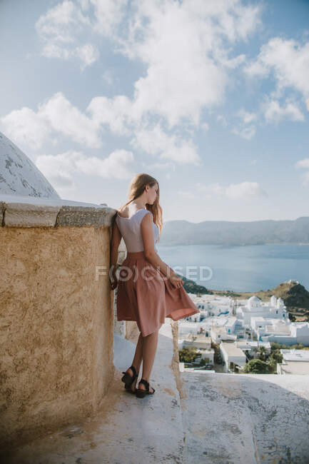 Vista trasera anónima delgado viajero femenino en vestido de fiesta de moda de pie en las escaleras de piedra de mala calidad en el pequeño pueblo costero con cabañas blancas en el tiempo soleado en Grecia - foto de stock