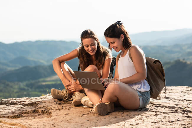 Захоплені подорожуючі друзі-жінки сидять на скелі в високогір'ї і дивляться фільм на нетбук під час літніх походів — стокове фото