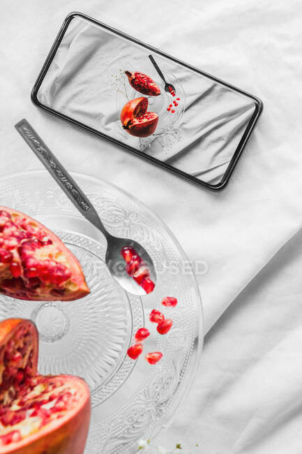 Top view of bright pomegranate seeds with spoon on stand near cellphone with photo on screen on white background — Stock Photo