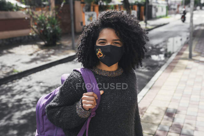 Side view of delighted ethnic female student with Afro hairstyle and backpack standing on street on sunny day with face mask and looking at camera — Photo de stock