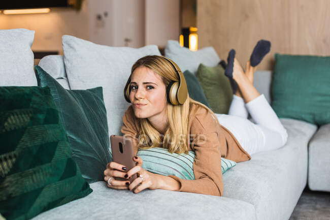 Радісна жінка лежить на дивані і насолоджується музикою в навушниках, кидаючись на камеру — стокове фото