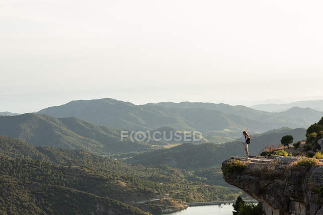 Vista remota del excursionista femenino de pie con los brazos extendidos en el borde de la colina y disfrutando de la libertad durante el trekking en las montañas en verano - foto de stock
