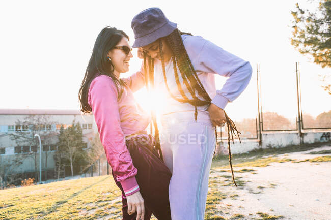 Smiling diverse lesbian girlfriends in trendy wear embracing and speaking with back light — Stock Photo
