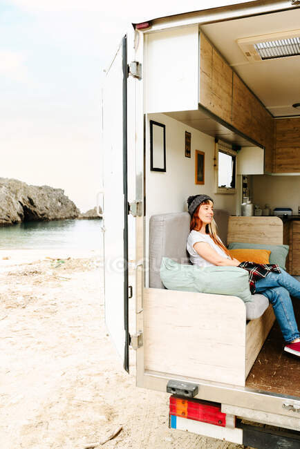 Vista lateral de la mujer hipster viajero feliz sentado en el sofá en el camión y disfrutar de vacaciones cerca del lago - foto de stock