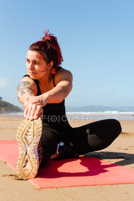 Deportiva adulta con tatuajes haciendo ejercicio en la alfombra mientras mira hacia otro lado contra el océano bajo el cielo azul - foto de stock
