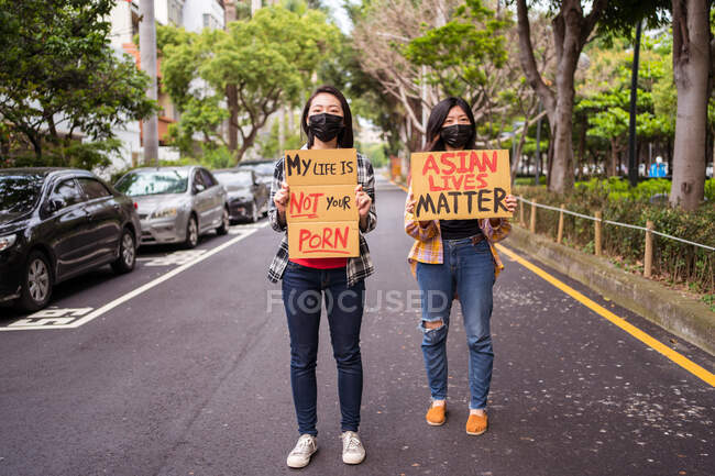 Mulheres étnicas em máscaras segurando cartazes protestando contra o racismo na rua da cidade e olhando para longe — Fotografia de Stock