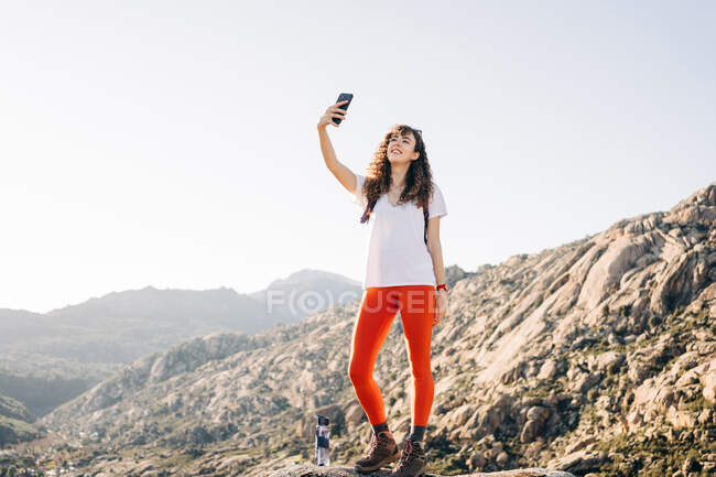 Corps complet de la jeune voyageuse positive avec les cheveux noirs bouclés dans des vêtements décontractés souriant tout en prenant selfie sur téléphone portable pendant la randonnée dans les montagnes par jour ensoleillé — Photo de stock