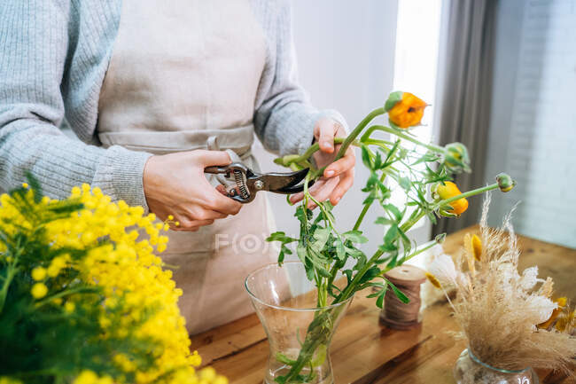 Crop unrecognizable young female florist in casual clothes cutting stems of fresh bright flowers with pruning snips while arranging bouquet in glass vase — Stock Photo