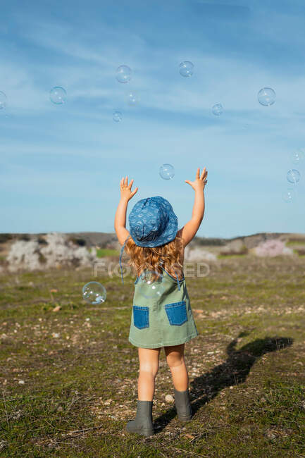 Back view of little girl in denim overalls playing with soap bubbles flying over green meadow on sunny day in countryside — Stock Photo