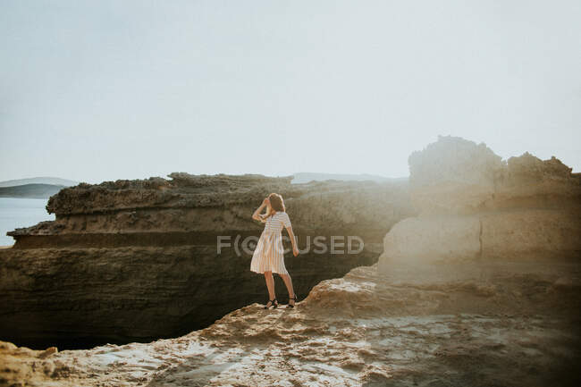 Full body anonymous fit female wearing white sundress standing gracefully on rough rocky cliff in seafront on sunny day in Sarakiniko Greece — Stock Photo