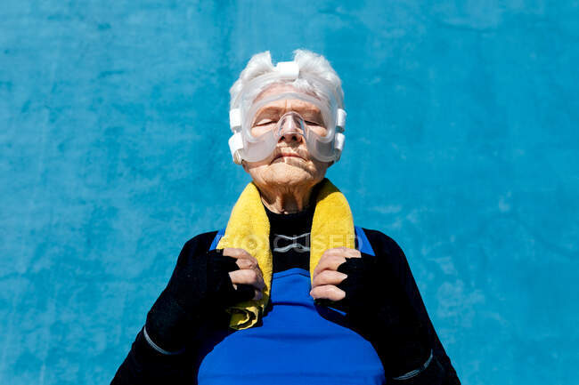 De baixo grave fêmea madura em activewear em protetor protetor de cabeça de boxe com toalha no ombro em pé no fundo azul com olhos fechados — Fotografia de Stock