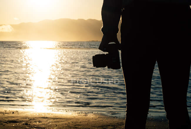 Back view silhouette of cropped unrecognizable traveler with photo camera standing contemplating seashore at sunset time - foto de stock