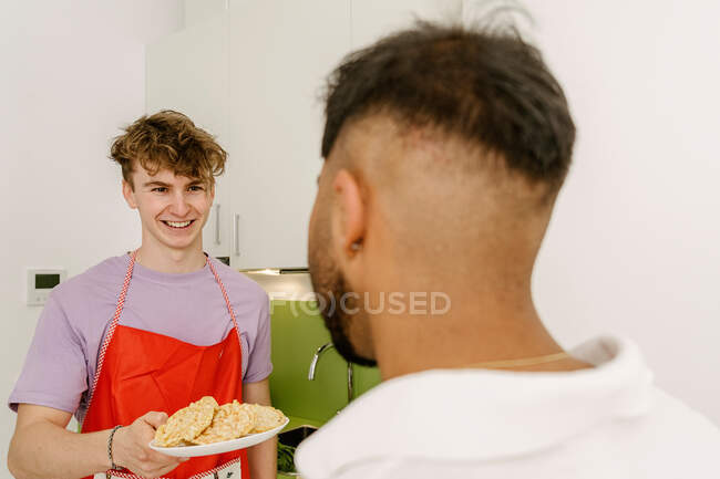 Cheerful young guy in casual clothes and apron giving plate of fresh prepared food to unrecognizable ethnic male friend in kitchen — Photo de stock