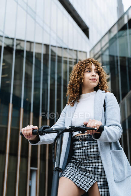 Young African American female in stylish wear standing with scooter on city street and looking away — Stock Photo