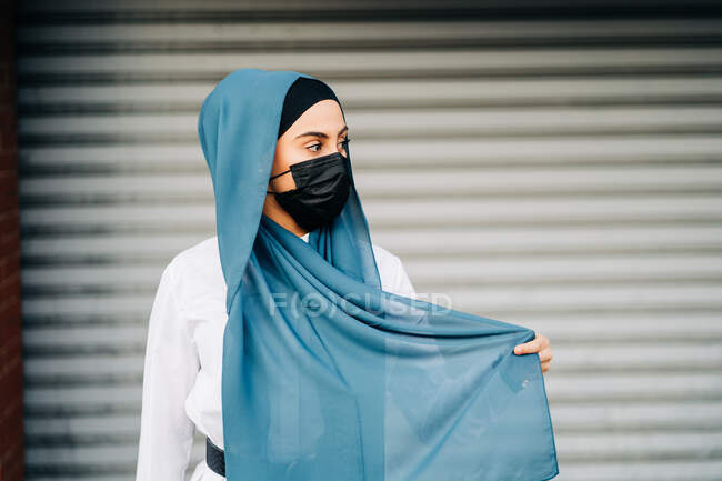 Mujer musulmana con máscara protectora y pañuelo tradicional de pie contra la pared en la ciudad y mirando hacia otro lado - foto de stock