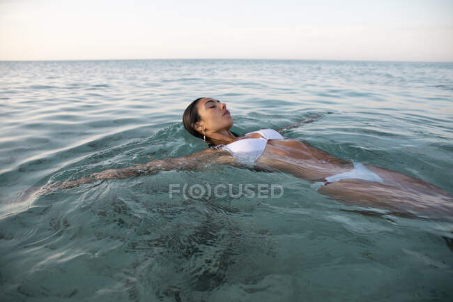 Mulher jovem em roupa de banho com braços estendidos e olhos fechados deitada no oceano com água transparente — Fotografia de Stock