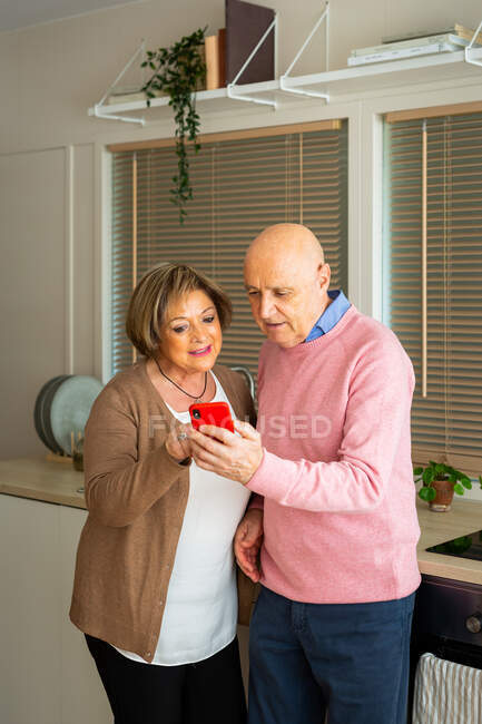 Middle aged man talking to woman using smartphone while standing together in kitchen at home — Stock Photo