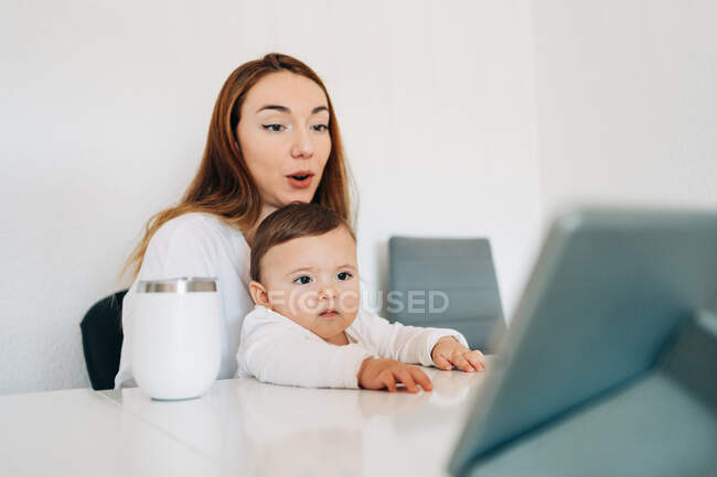 Positiva joven madre y lindo bebé atento viendo dibujos animados en la tableta mientras están sentados en el escritorio juntos - foto de stock