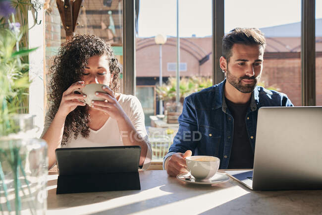 Funcionário remoto feminino étnico desfrutando de bebida quente contra tablet e parceiro masculino com laptop à mesa à luz do sol — Fotografia de Stock
